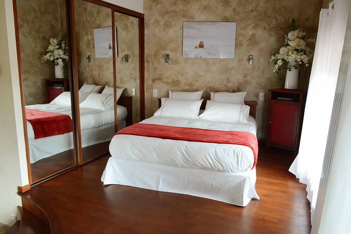 Uma cama de casal com um cobertor vermelho, piso de madeira, um espelho, um quadro na parede e um vaso com flores.