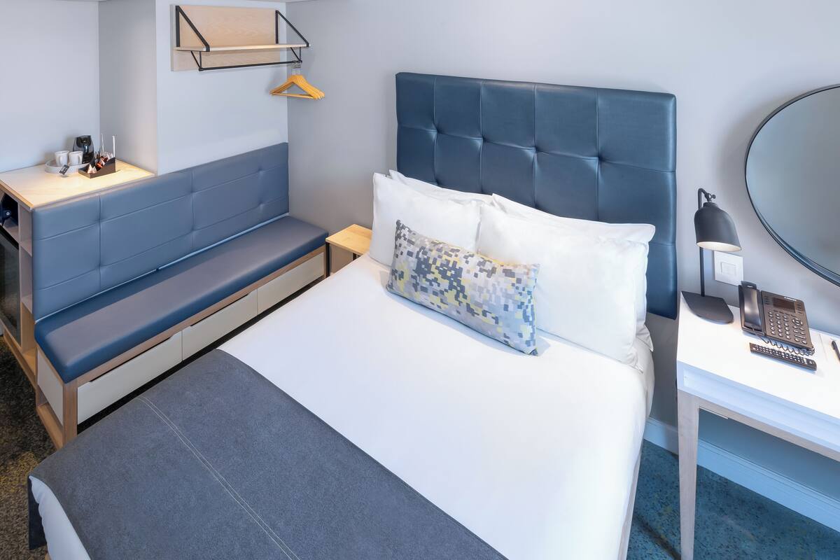 Une chambre d’hôtel avec un lit, une table de chevet avec une lampe, une petite table avec un téléphone et un banc.
