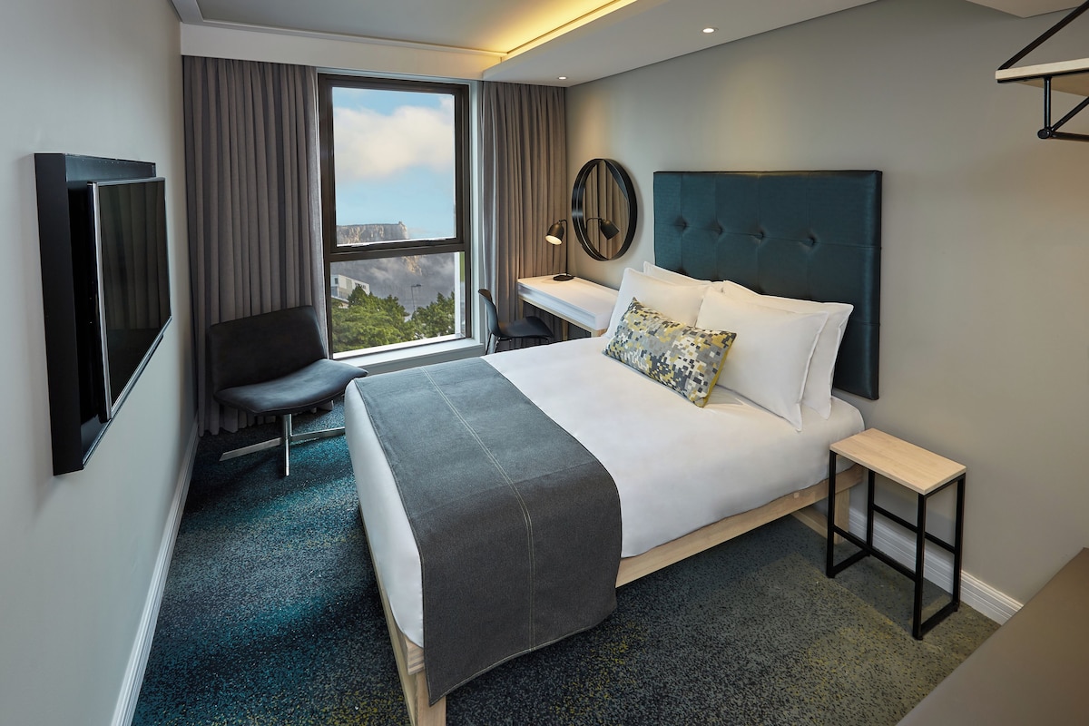 Une chambre d’hôtel moderne équipée d’un lit, d’un bureau, d’une chaise, d’une télévision et offrant une vue sur l’extérieur.