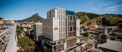 Un hotel moderno con un llamativo letrero 'Stayasy', rodeado de edificios urbanos y un paisaje montañoso.