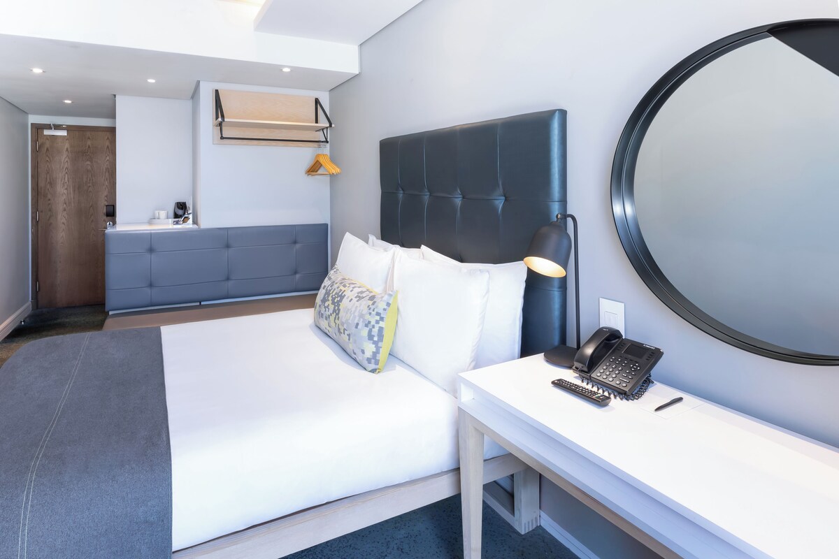 A modern hotel room with a large bed, a bedside table with a phone, a round mirror, and a small desk.