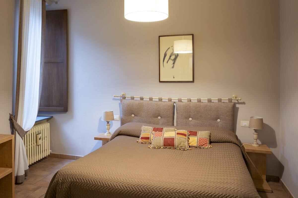 Een slaapkamer met een groot bed, een hoofdbord met knopen, twee nachtkastjes met lampen, een radiator en een raam met gordijnen.