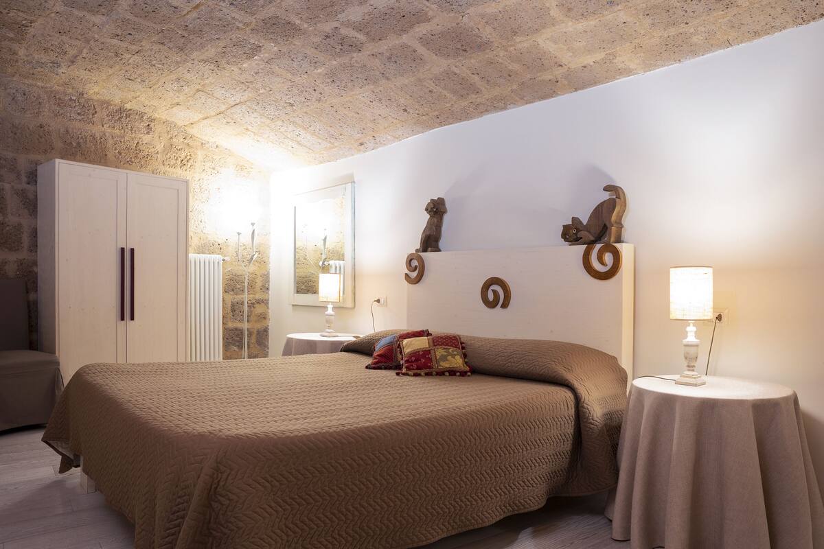 Een slaapkamer met een groot bed, een kledingkast, een rond tafeltje met een lamp en decoratieve wandkunst.