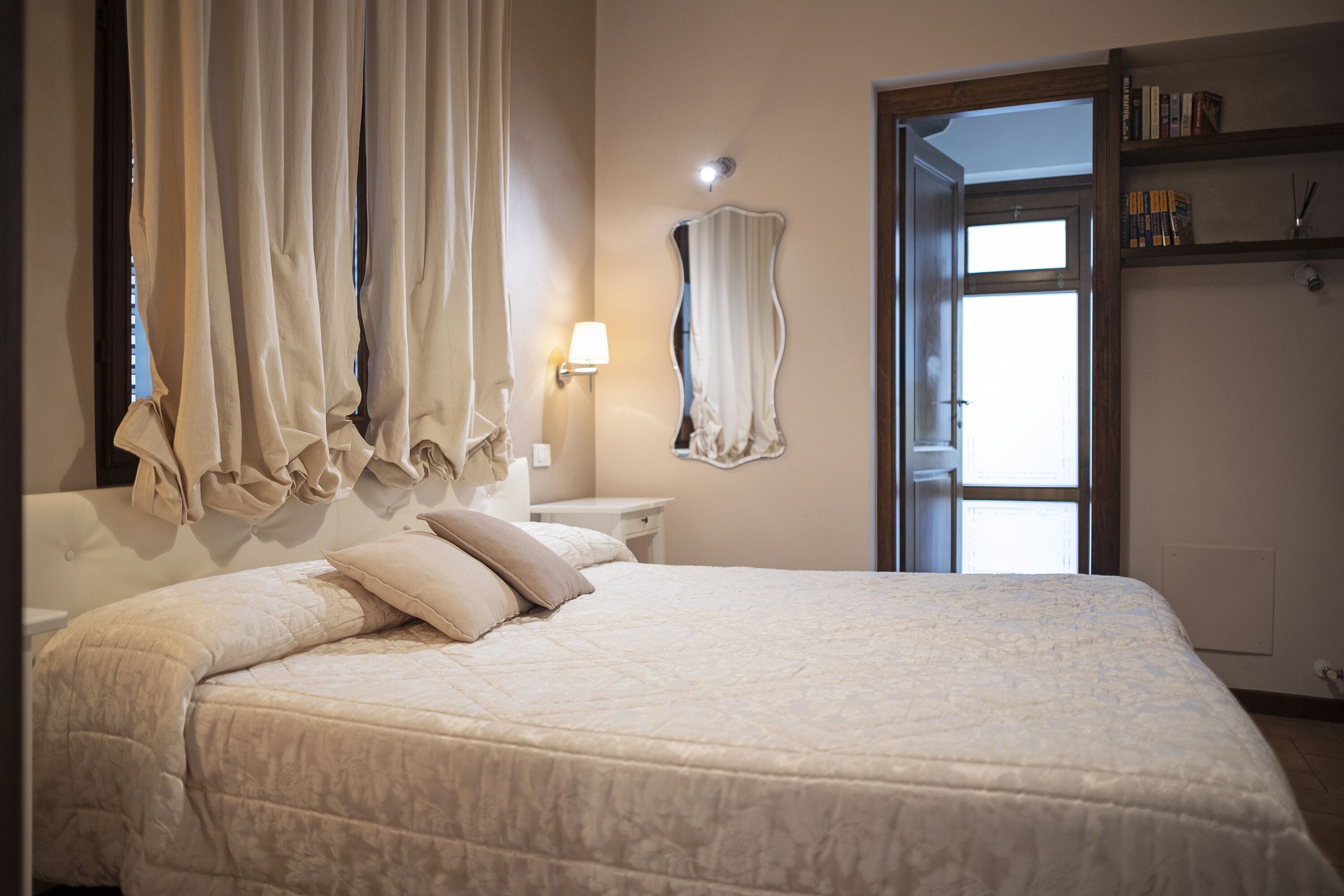 A bedroom with a large bed, a bedside table, a lamp, a mirror, and a bookshelf.