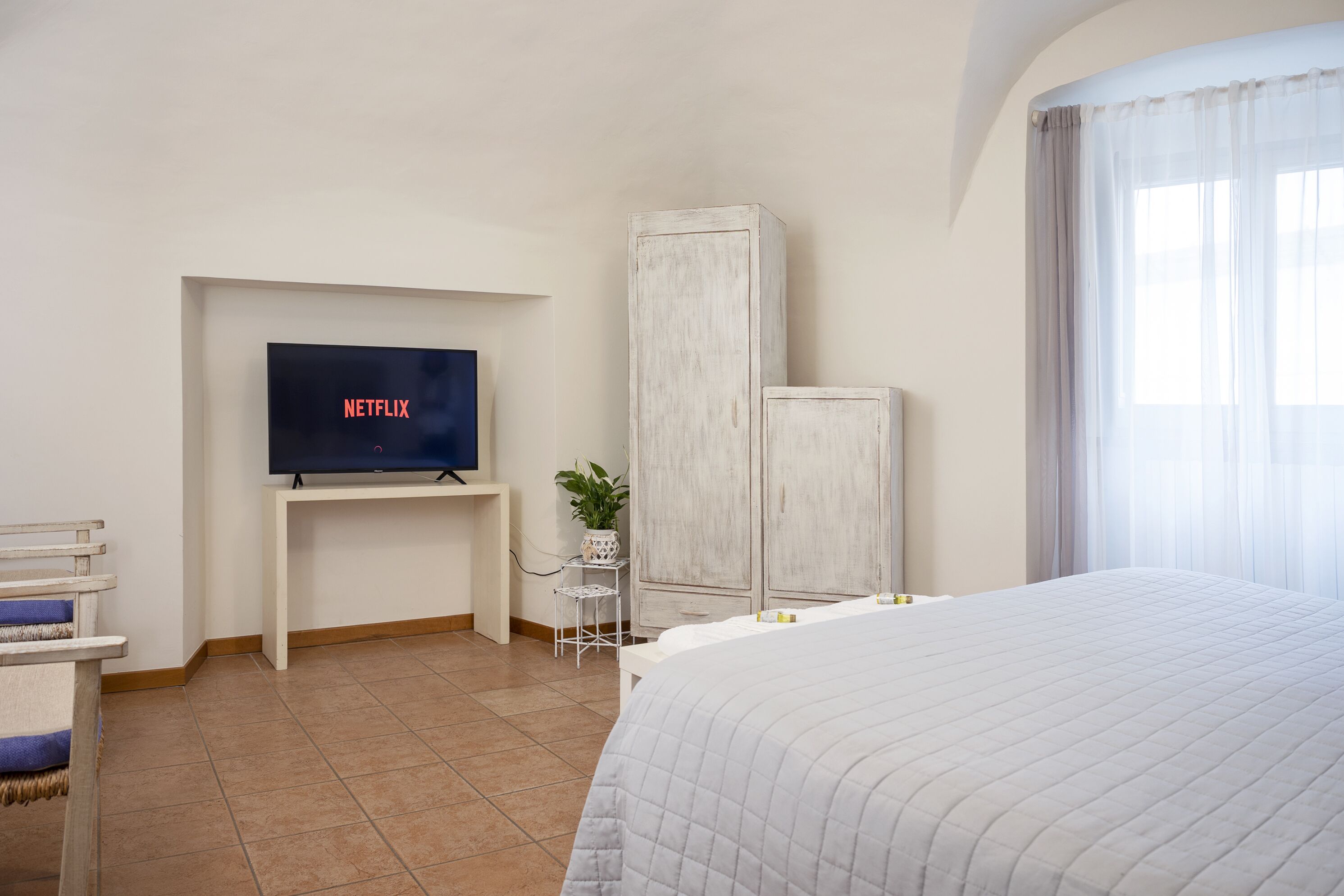 Una camera d'hotel con un letto, una televisione su un mobile, un armadio, un tavolino con una pianta e una finestra con tende.