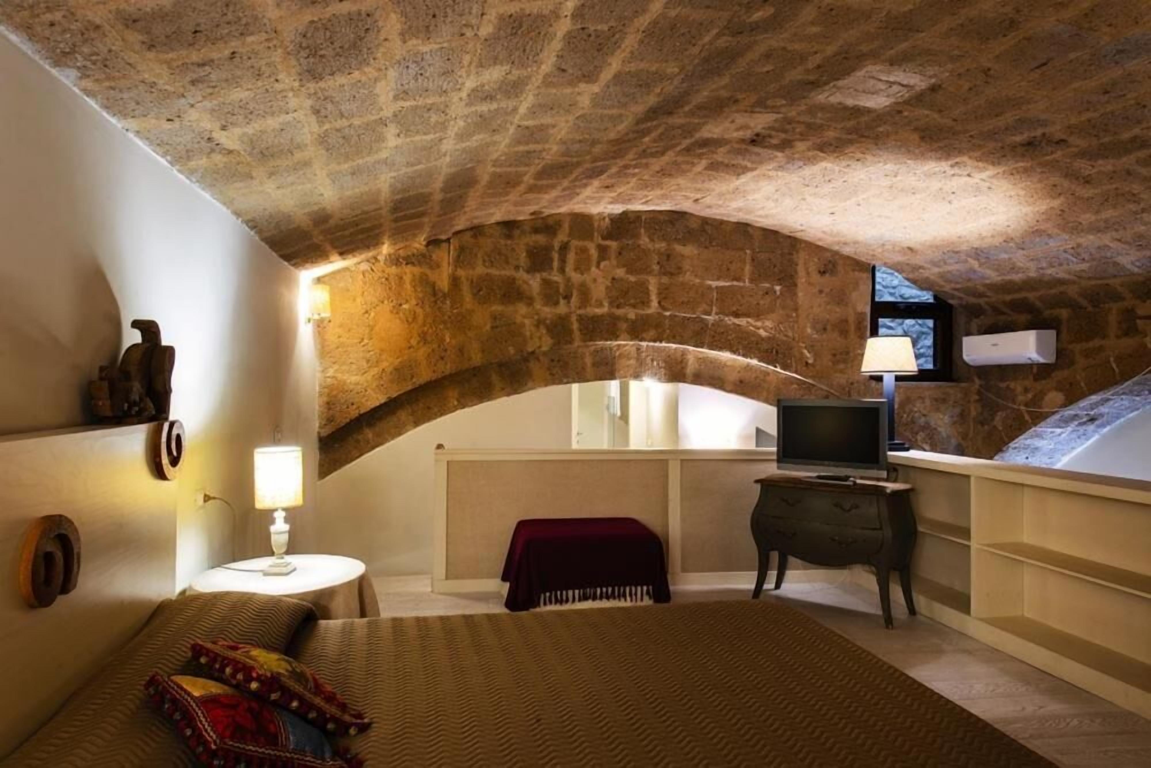 Un dormitorio con techo de piedra, una cama con manta roja, una mesita de noche con lámpara, un televisor sobre un soporte y una ventana con vistas.