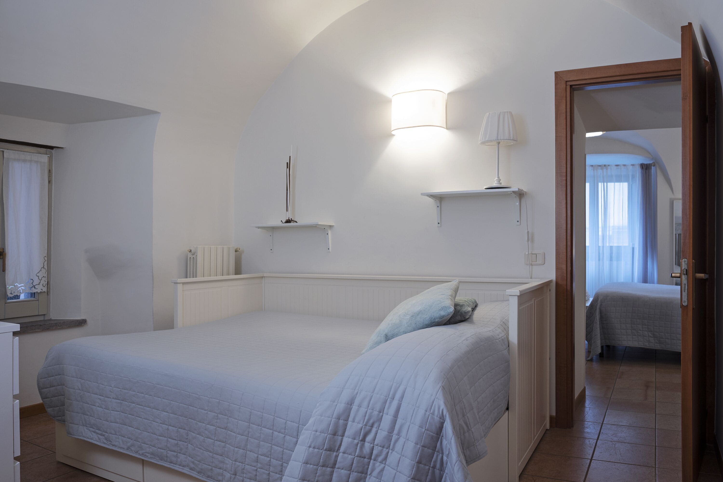 Een slaapkamer met een groot bed, een nachtkastje, een wandlamp en een deur naar een andere kamer.