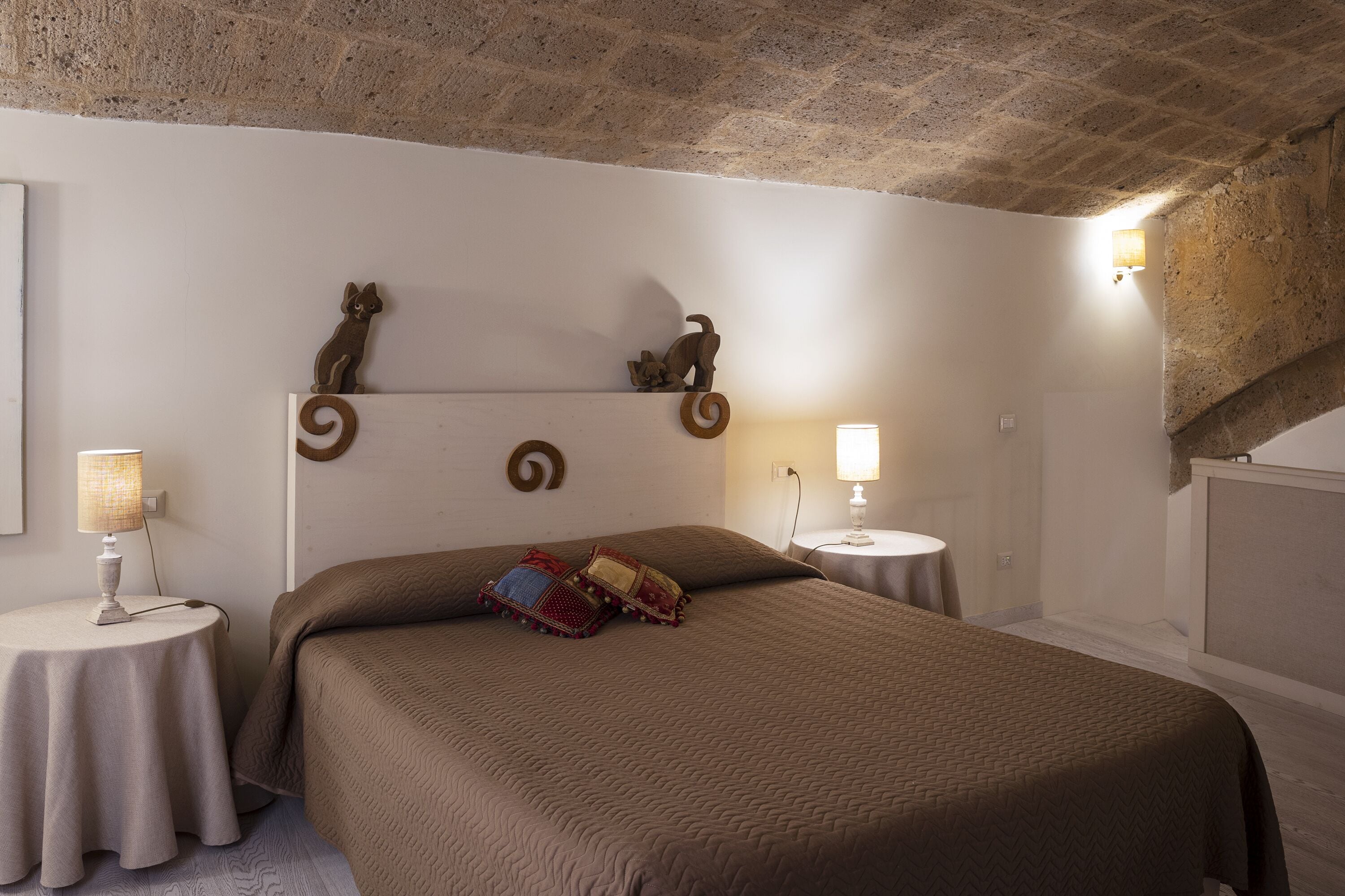 Une chambre avec un plafond en briques, un grand lit recouvert d’une couette marron, deux tables de chevet avec des lampes et des éléments de décoration fixés au mur.