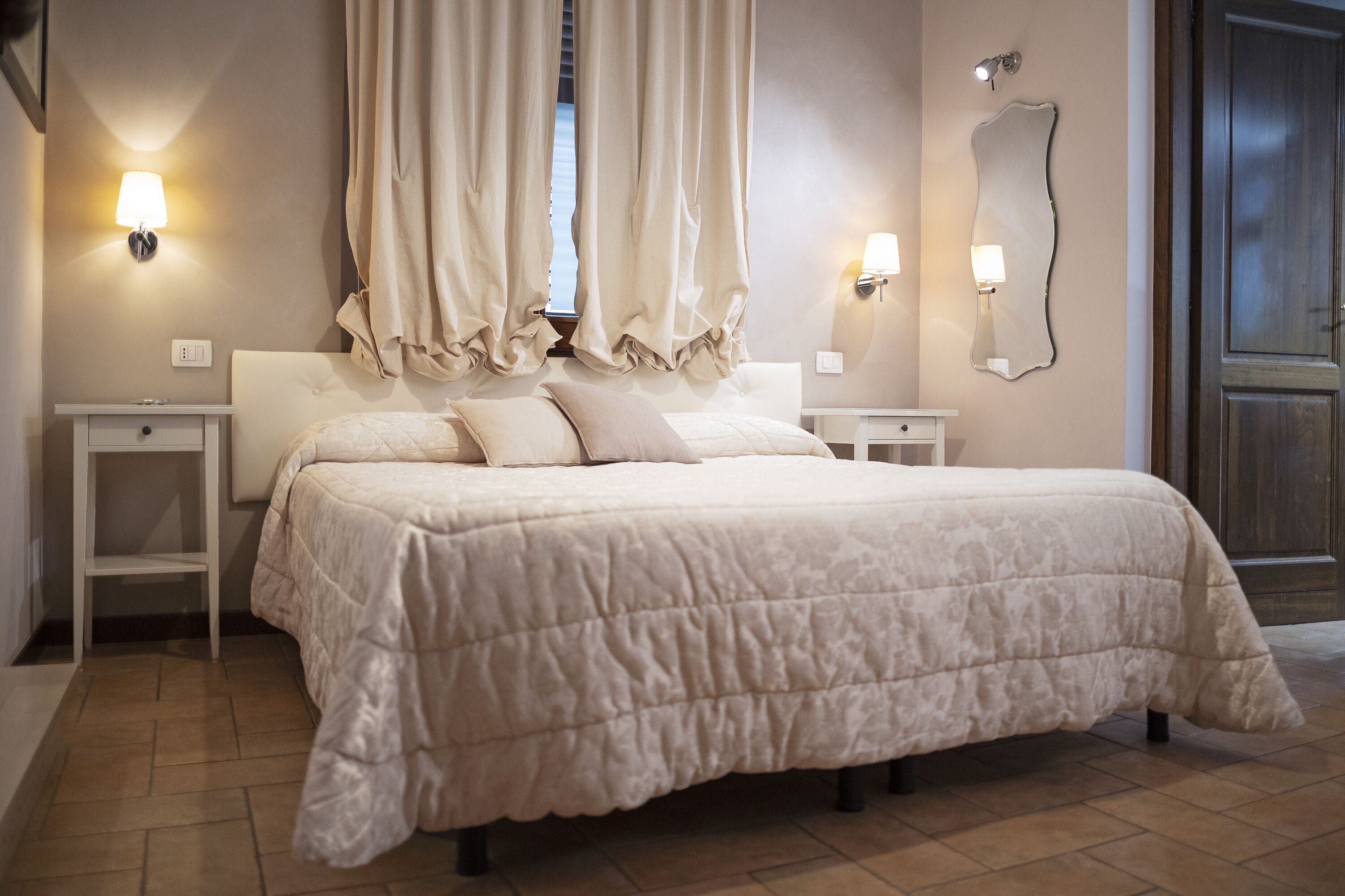 A bedroom with a large bed, two bedside tables, a wall-mounted mirror, and a window with curtains.