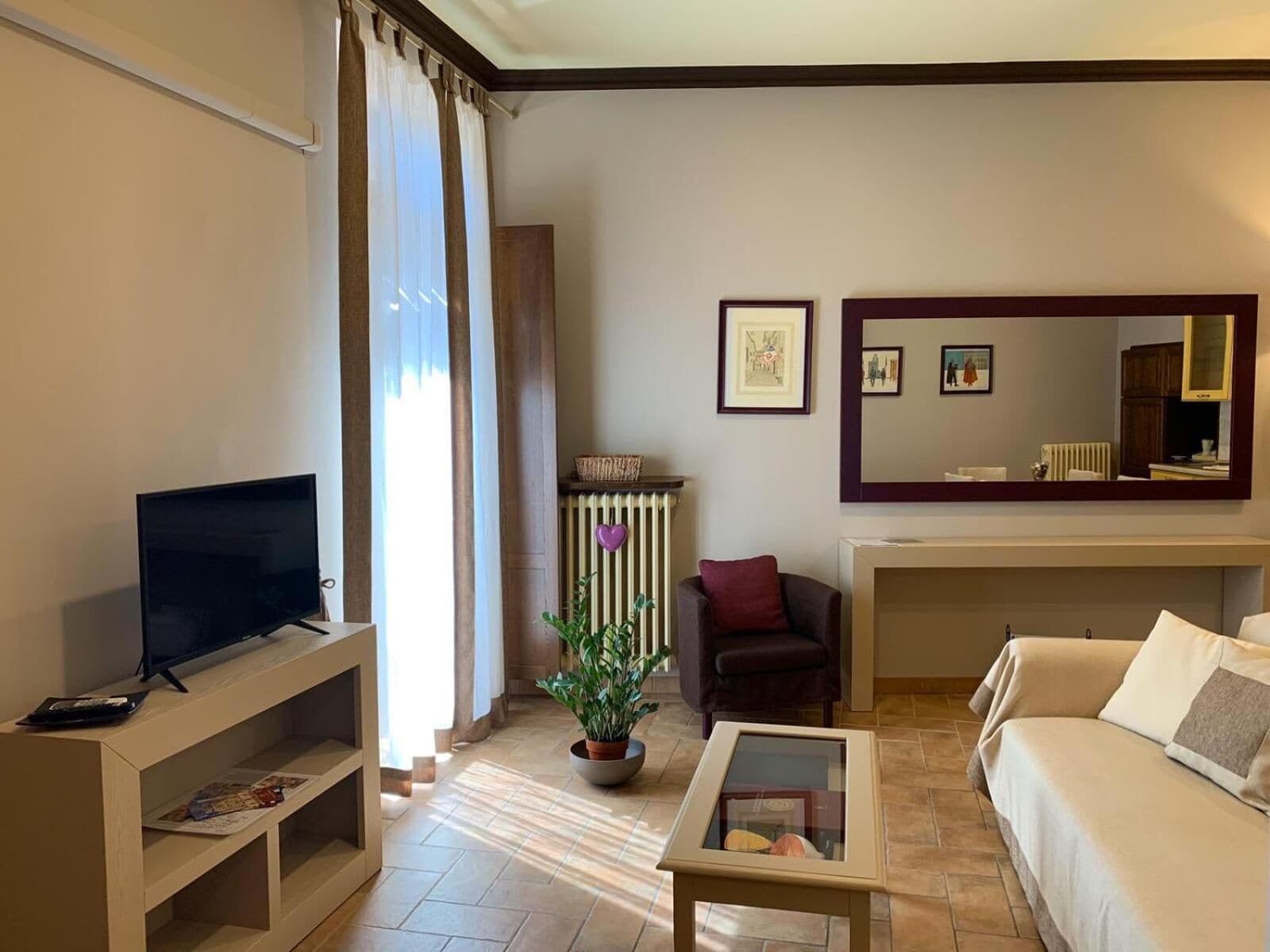 Habitación de hotel con televisión sobre un mueble bajo, sofá, una mesita, planta en maceta y un espejo grande.