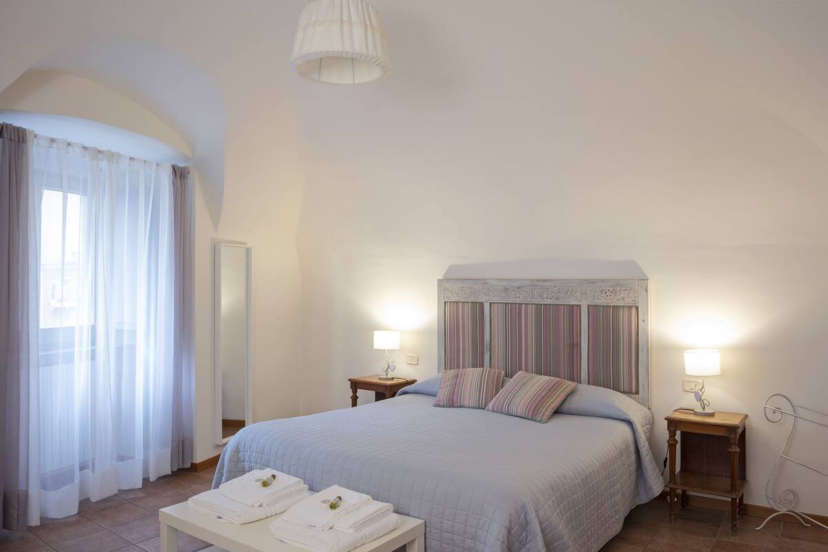 Een netjes ingerichte slaapkamer met een groot bed, twee nachtkastjes met lampen, een klein tafeltje met opgevouwen handdoeken en een raam met dunne gordijnen.