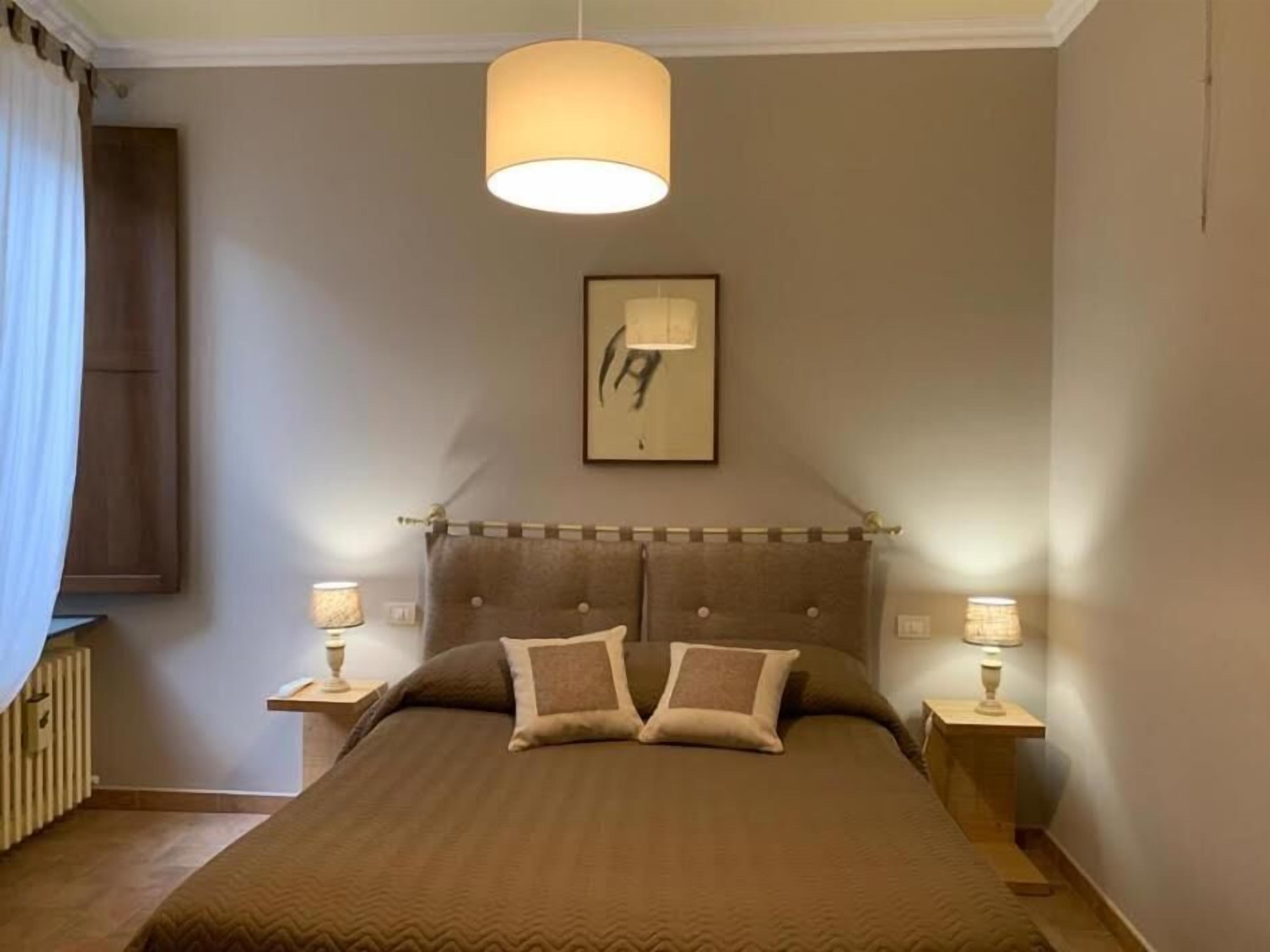 Een slaapkamer met een groot bed, twee bedlampjes, een plafondlamp, een lijstwerk en een raam met gordijnen.