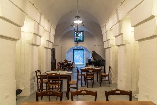 Un comedor con varias mesas y sillas, un piano y un amplio ventanal arqueado.