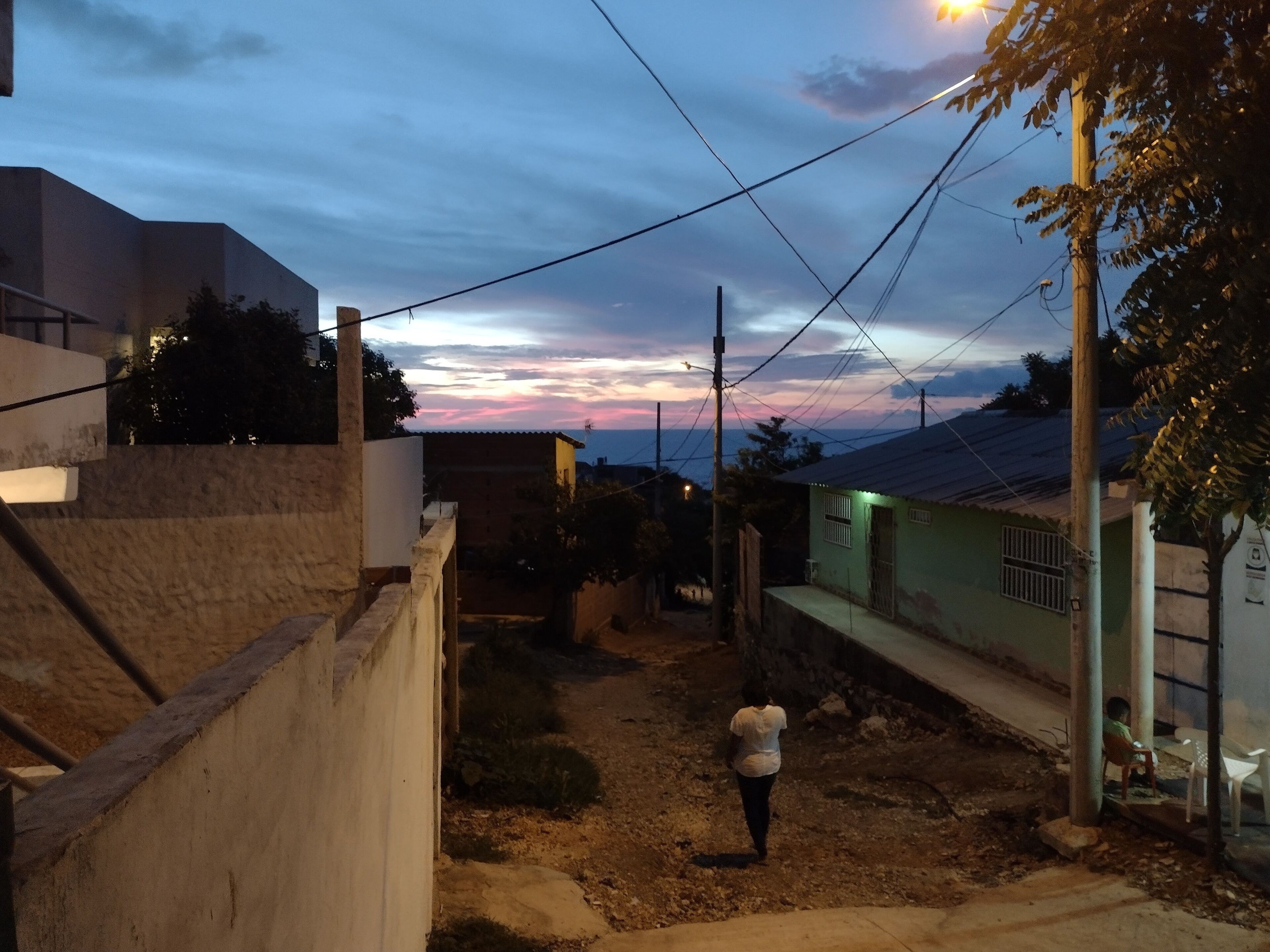 Um beco estreito com uma pessoa se afastando da câmera, cercado por edifícios com janelas e portas visíveis.