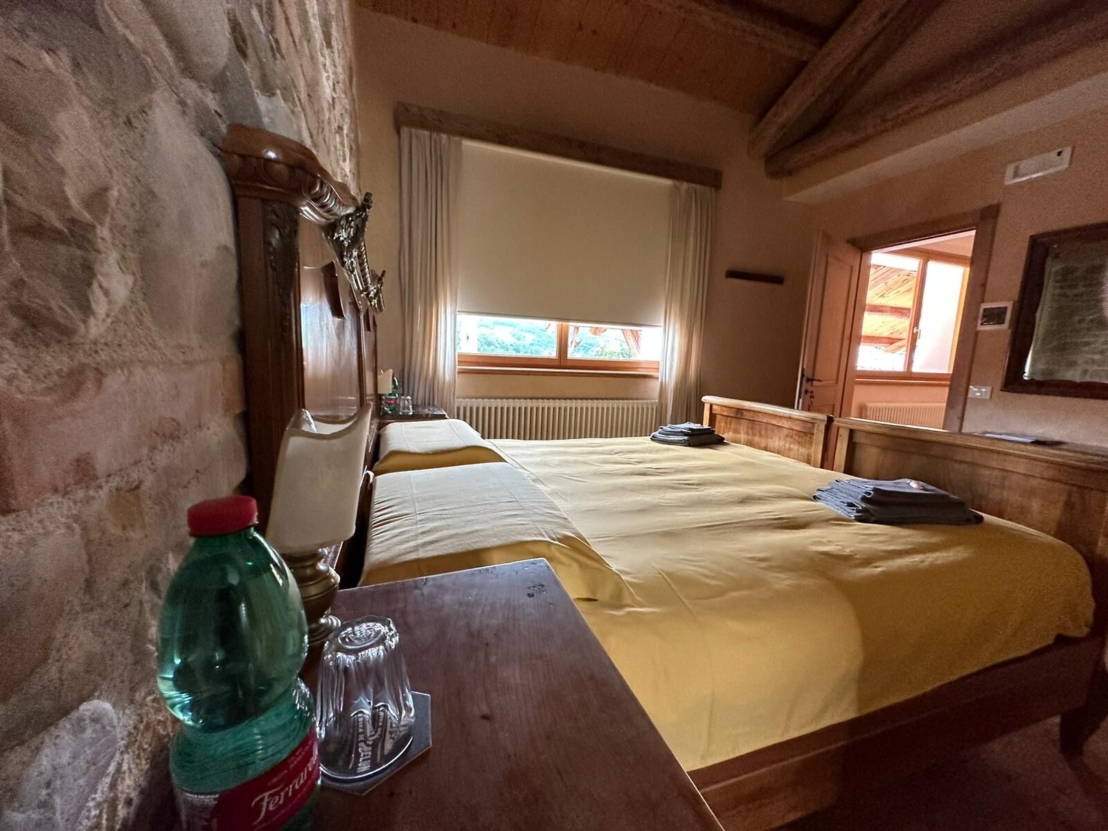 Una habitación con una cama de madera, una mesa de madera con una botella y un vaso, una ventana con cortinas y un radiador.