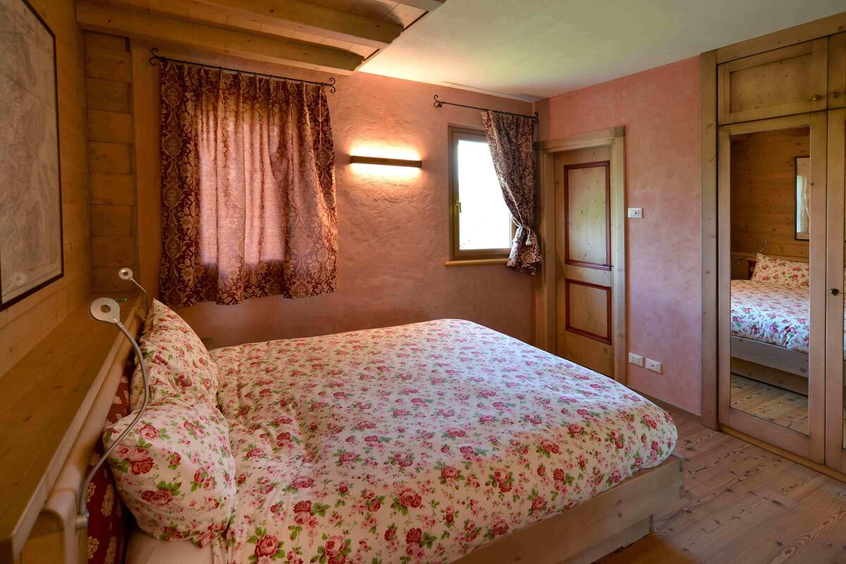 A bedroom with a wooden bed, floral bedding, a window with curtains, and a wooden wardrobe.