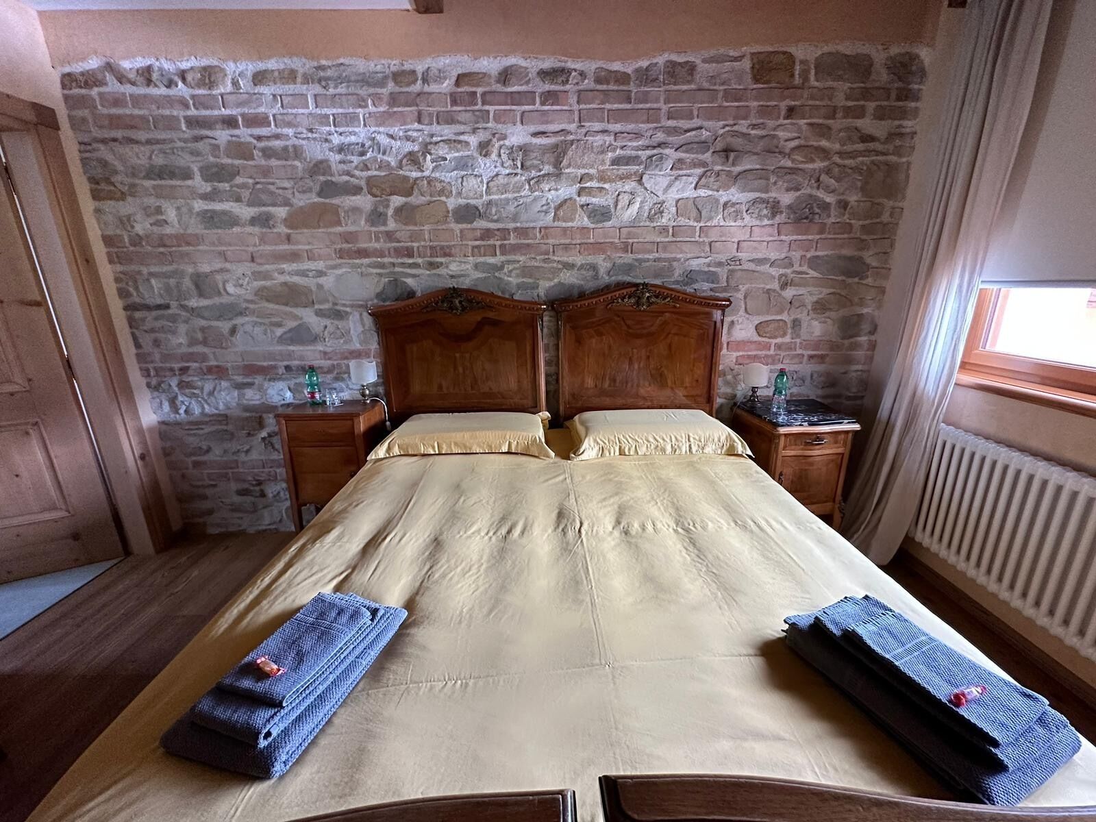 A double bed with a stone wall backdrop, wooden headboard, bedside tables with lamps, and a window with curtains.