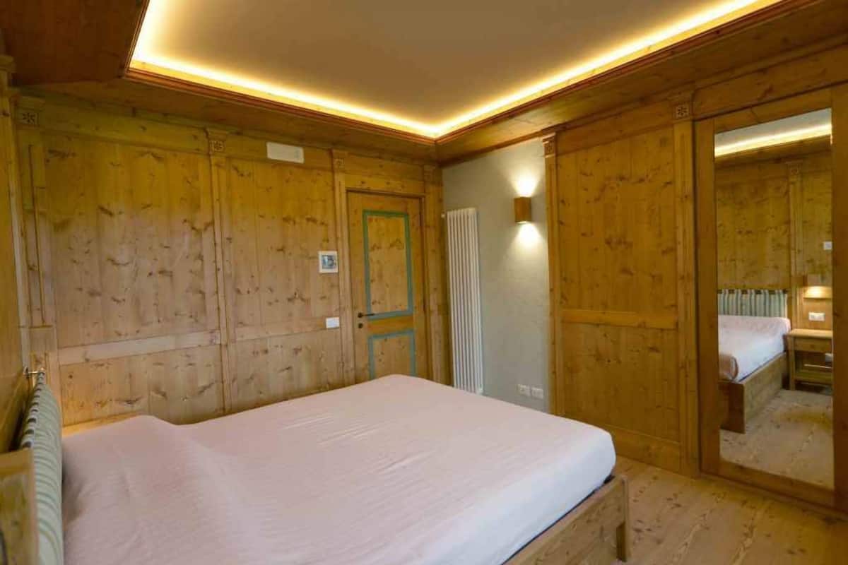 A wooden-paneled room with a bed, a radiator, and a mirror reflecting the room.