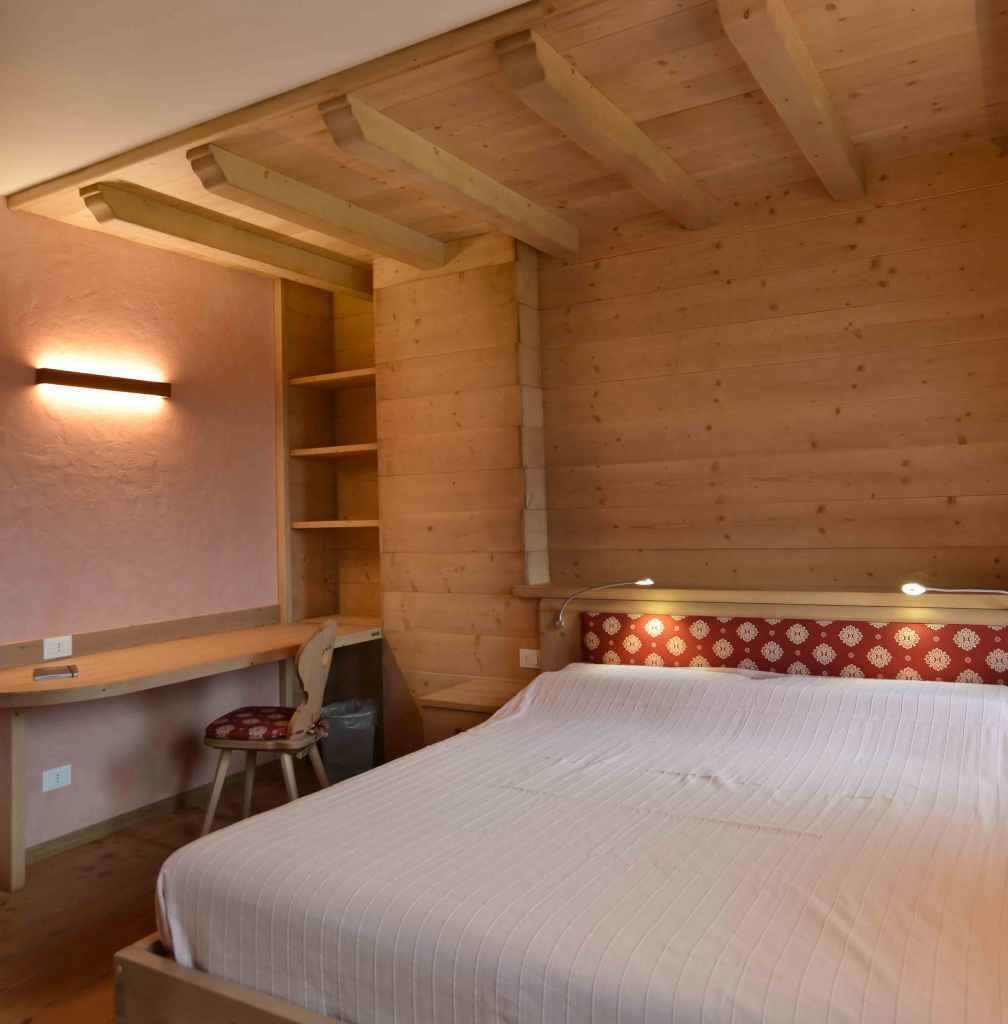 A cozy bedroom with a wooden ceiling, a bed with a patterned headboard, a desk with a chair, and a wall-mounted shelf.