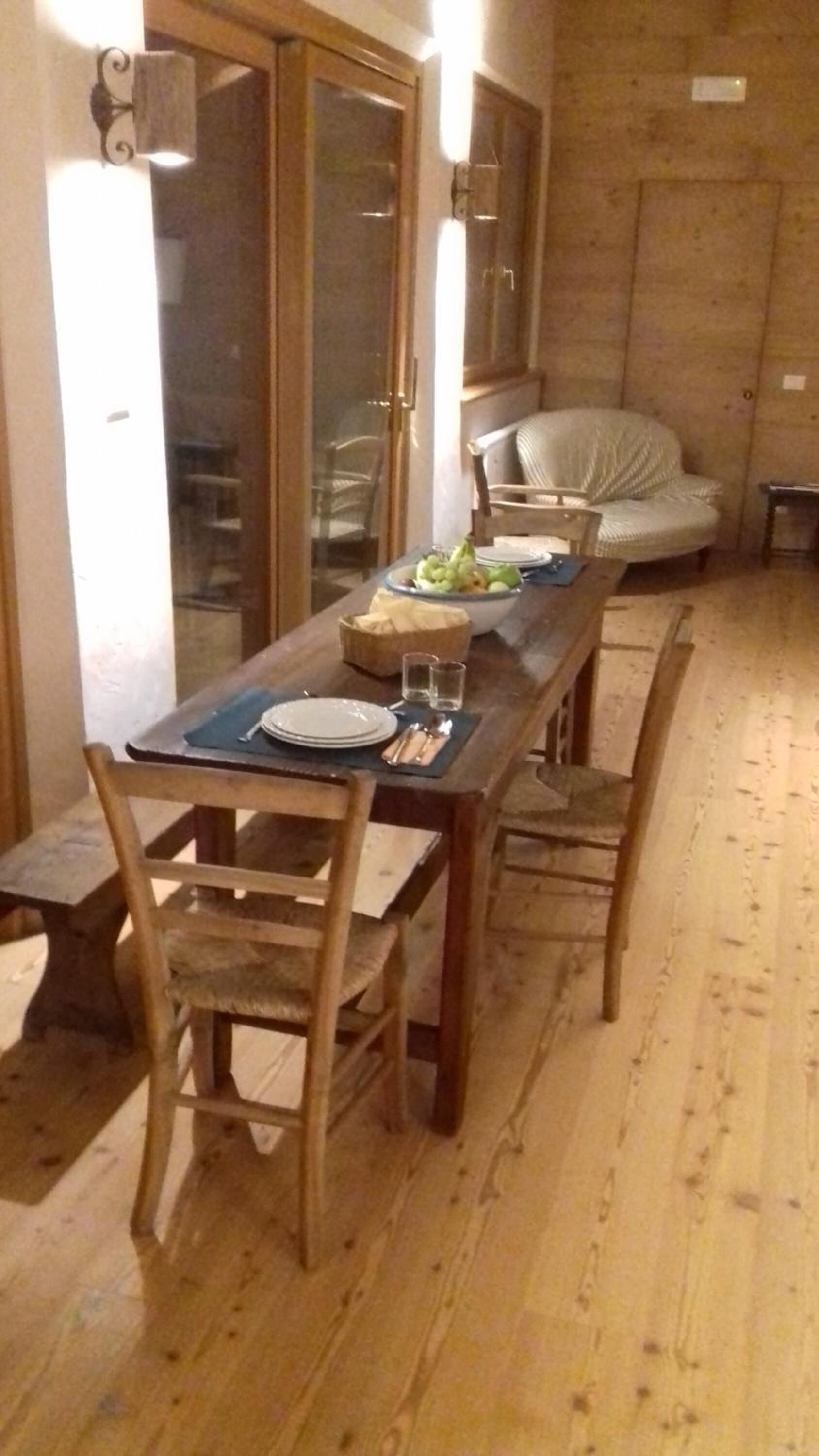 Un comedor con una mesa de madera dispuesta para dos personas, sillas y un bol de fruta.