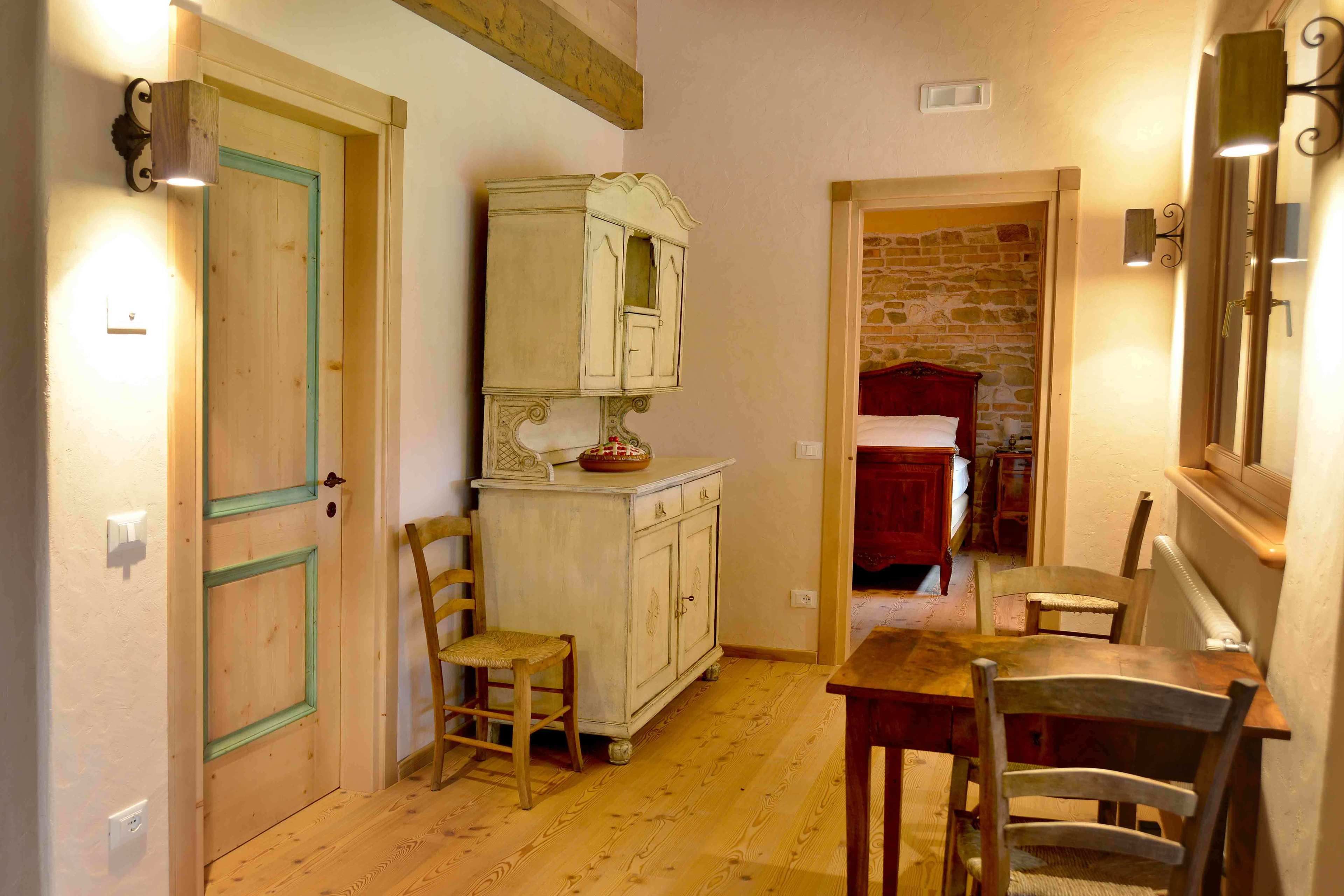 Un corridoio stretto con pavimento in legno, una porta in legno con stipite verde, un armadietto bianco con specchio e un letto rosso sullo sfondo.