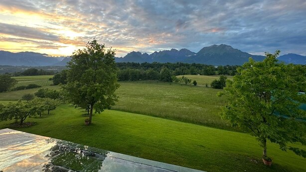 Une vue d’un paysage montagneux avec un champ herbeux et des arbres, sous un ciel nuageux.