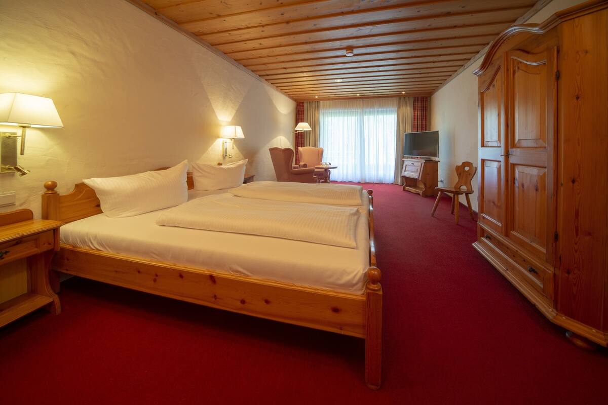 A hotel room with a wooden bed, two bedside tables, a chair, a TV, and a wardrobe.