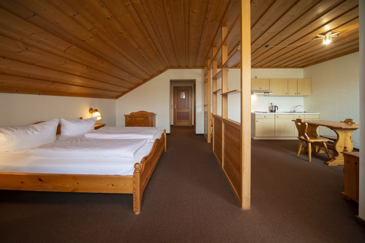 A bedroom with two wooden beds, a wooden ceiling, a carpeted floor, a wooden desk, and a kitchen area with wooden cabinets and a sink.