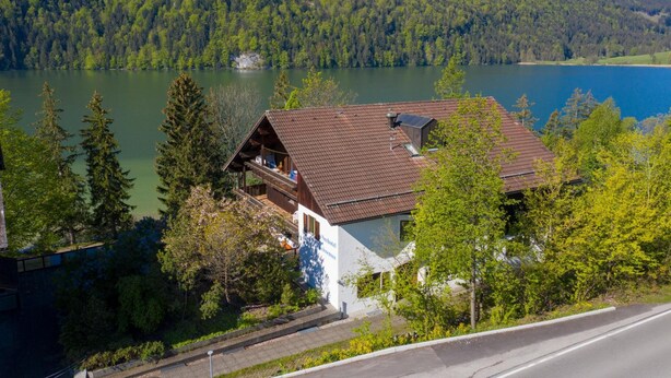 A chalet-style building with a sloped roof, situated by a lake with forested hills in the background. - Seehotel Weissensee (Fuessen)