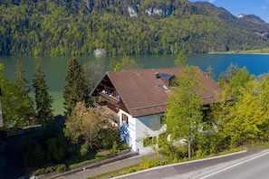 Um chalé com telhado inclinado, localizado à beira de um lago, com colinas arborizadas ao fundo.