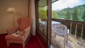 A room with a red carpet, a checkered sofa, a table with a magazine, a white chair, and a balcony with a view of a lake and mountains. - Seehotel Weissensee (Fuessen)