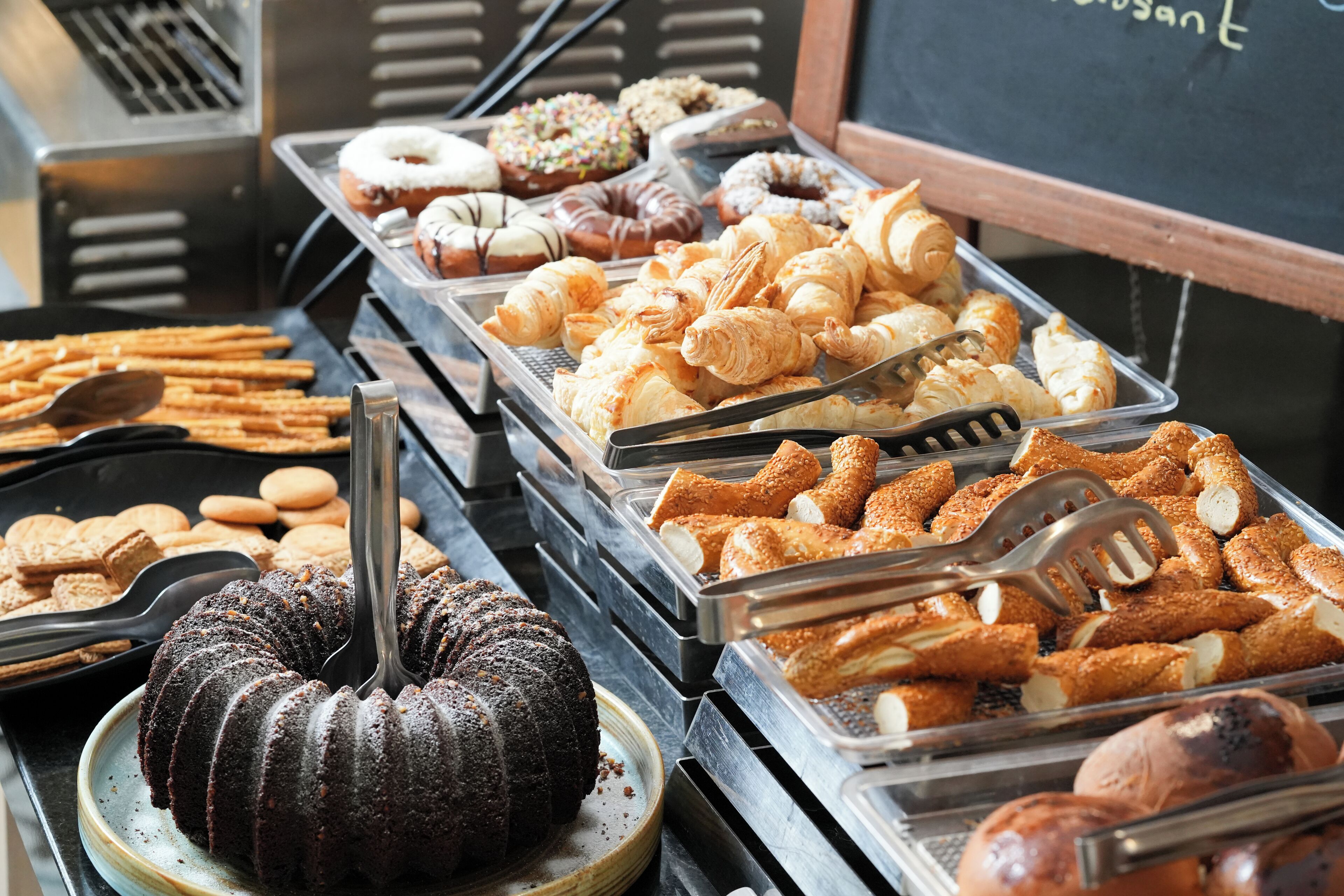 A variety of baked goods including donuts, croissants, and a bundt cake displayed in trays.