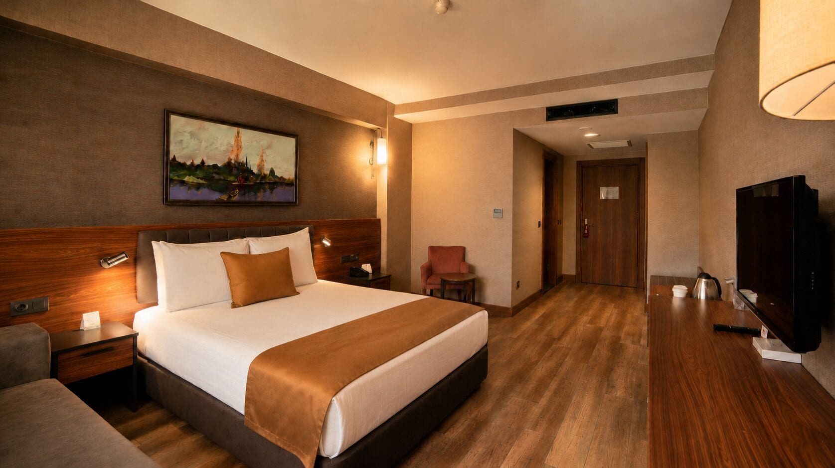 A hotel room with a bed, bedside table, lamp, chair, TV, and a painting on the wall.