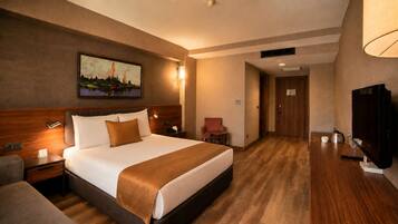 A hotel room with a bed, bedside table, lamp, chair, TV, and a painting on the wall.