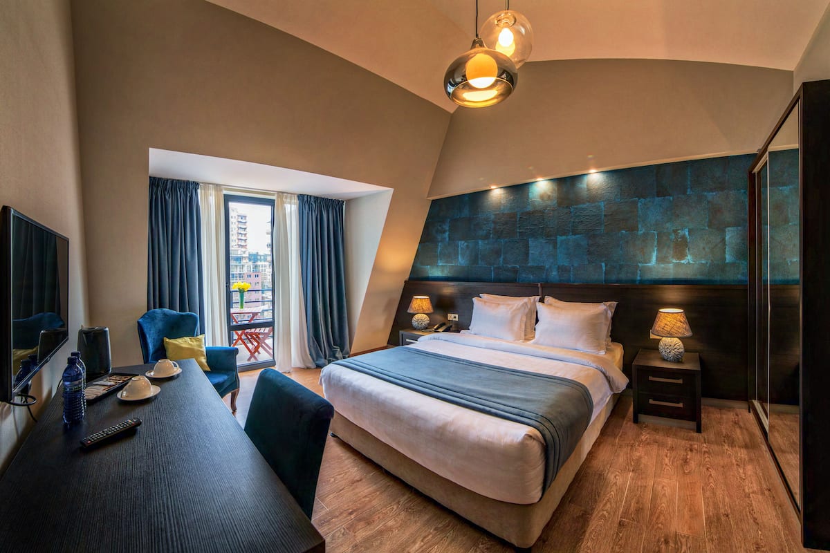 A modern hotel room with a large bed, a desk with a chair, a TV, and a balcony with curtains.