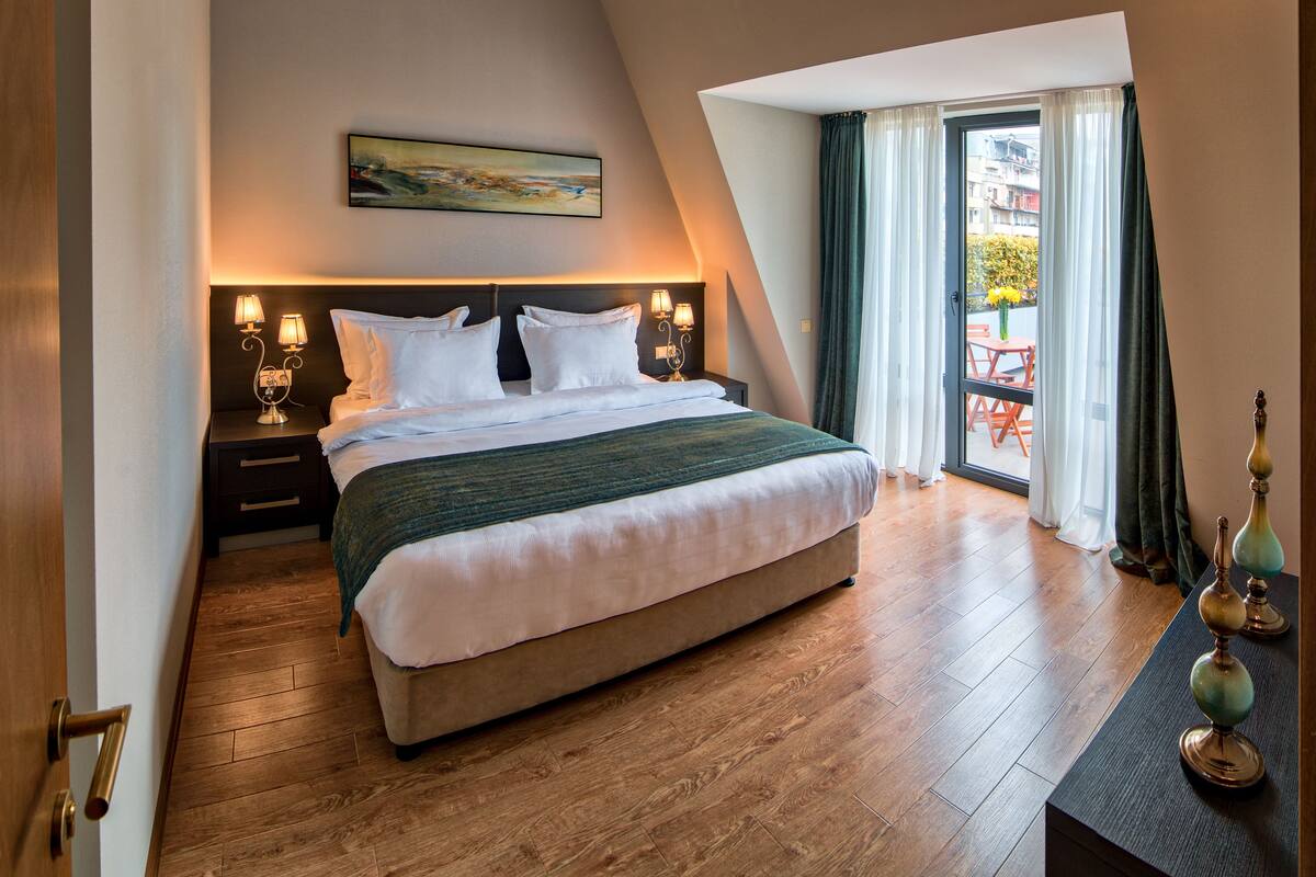 A bedroom with a large bed, wooden flooring, a painting on the wall, and a view through a window.