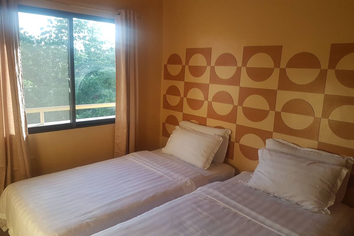 Huoneessa on kaksi sänkyä, ikkuna, jossa on verhot, ja seinä, jossa on geometrinen kuvio.