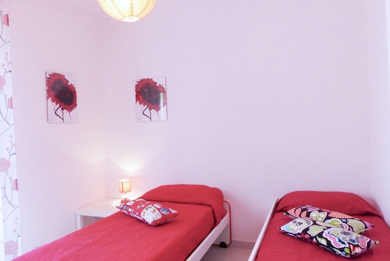 A bedroom with two single beds, each with a red bedspread and patterned pillows. There is a bedside table with a lamp and a small decorative item. The walls are plain with two abstract paintings.