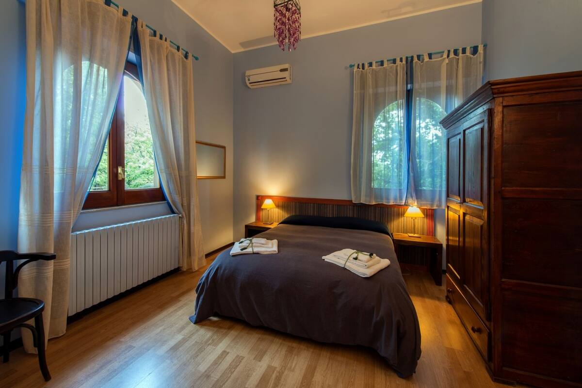 A hotel room with a large bed, two bedside lamps, a wooden wardrobe, and a window with curtains.