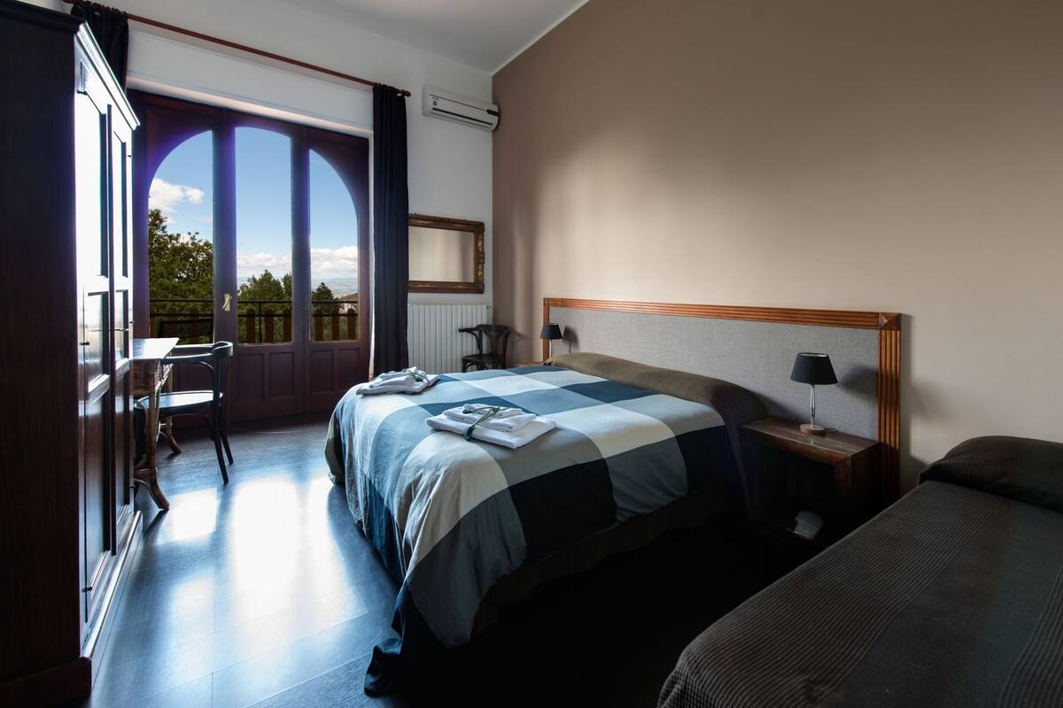 A hotel room with a large bed, a desk, and a chair. There is a window with a view of trees and a building.