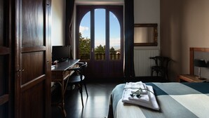 Een hotelkamer met een groot rondboogvenster, een bureau met stoel, een bed met opgemaakt bed en een spiegel aan de muur.