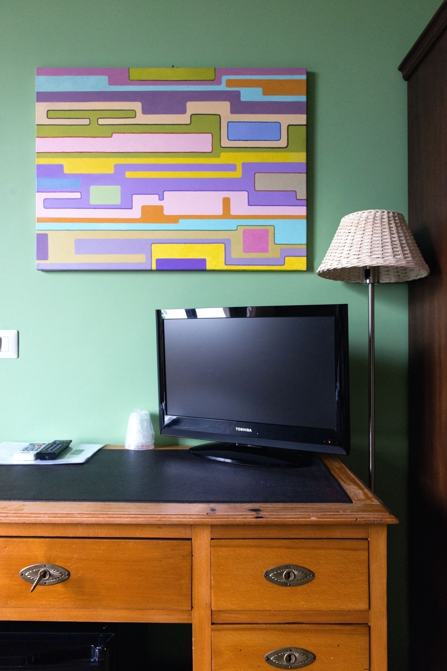 A room with a wooden desk, a flat-screen TV, a colorful abstract painting, and a floor lamp.