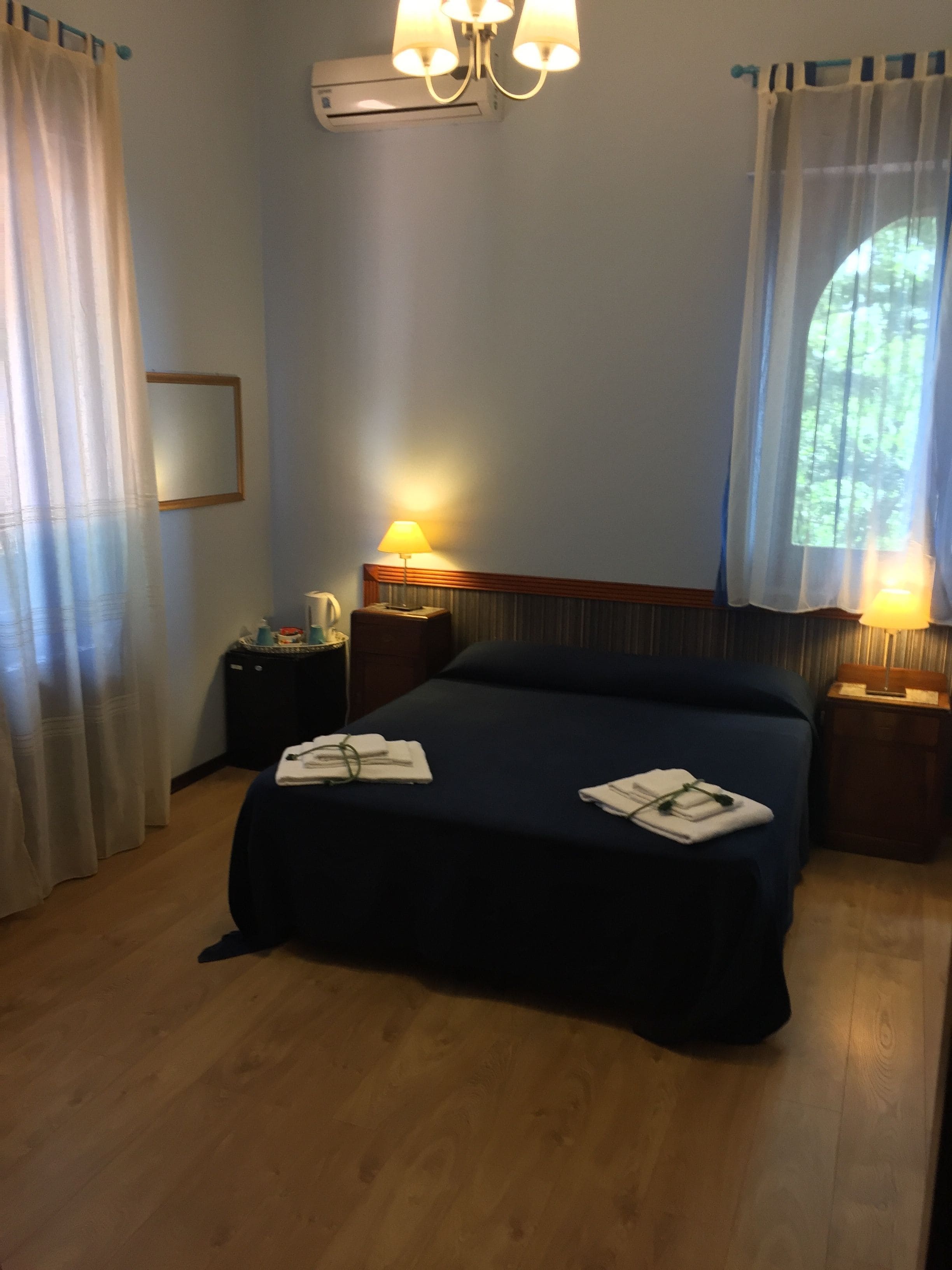 Una camera d'albergo con un letto, due comodini con lampade, una finestra con tende e un condizionatore d'aria a parete.