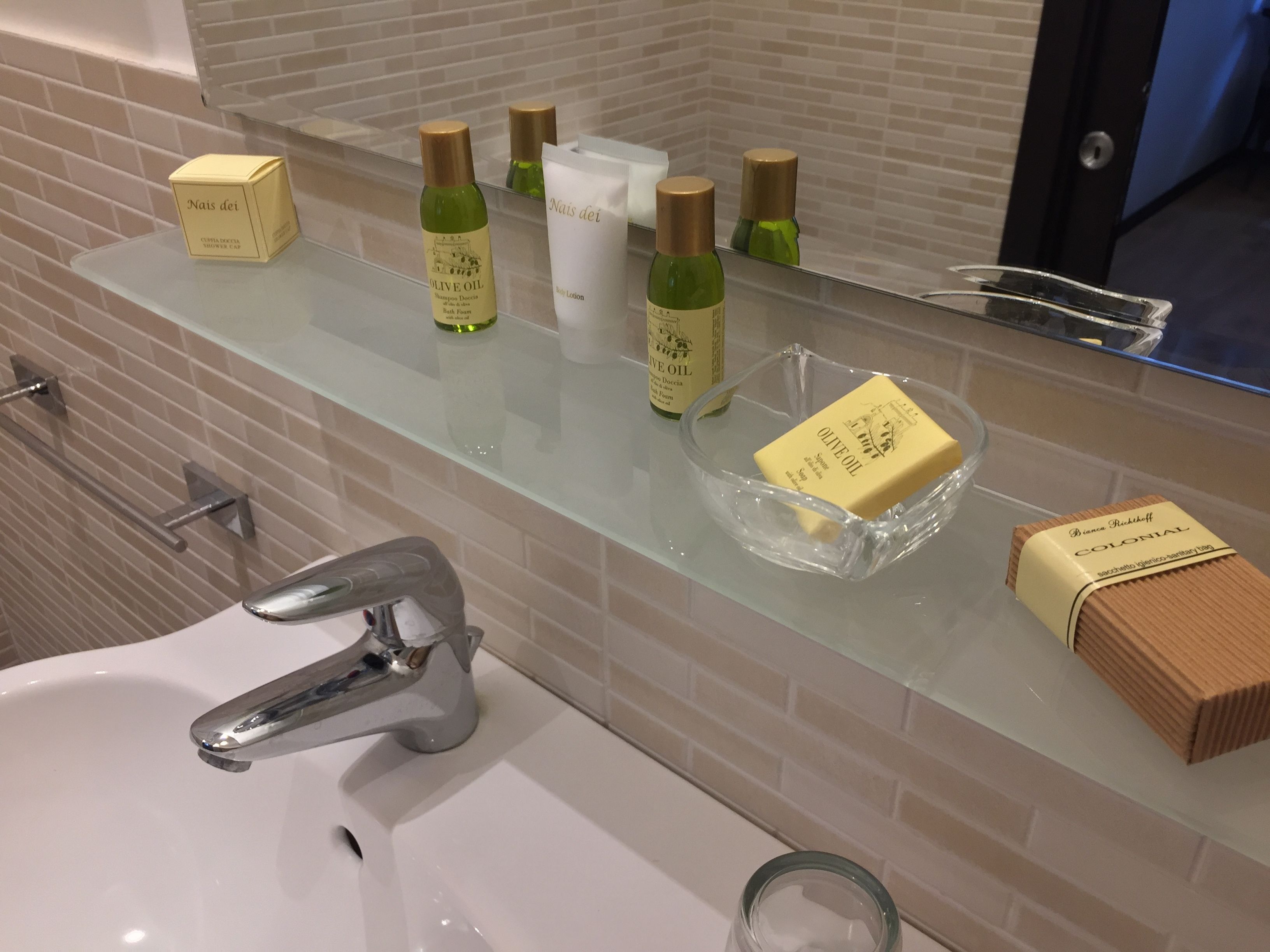 Un baño con repisas de vidrio que sostienen artículos de aseo, un lavamanos y un espejo.