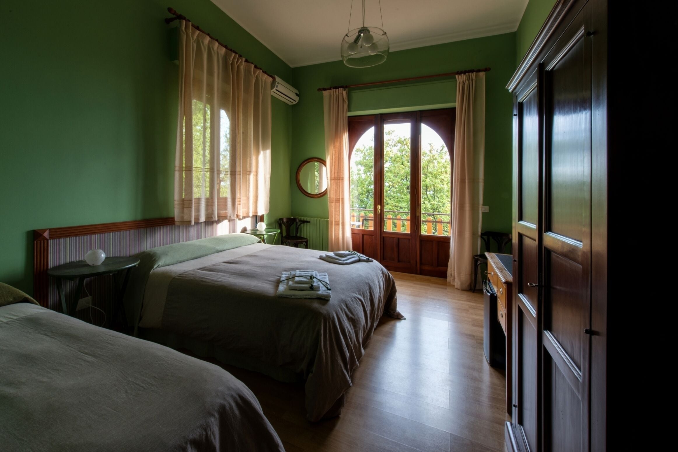 A room with two beds, a wooden floor, a large window with curtains, and a wardrobe.