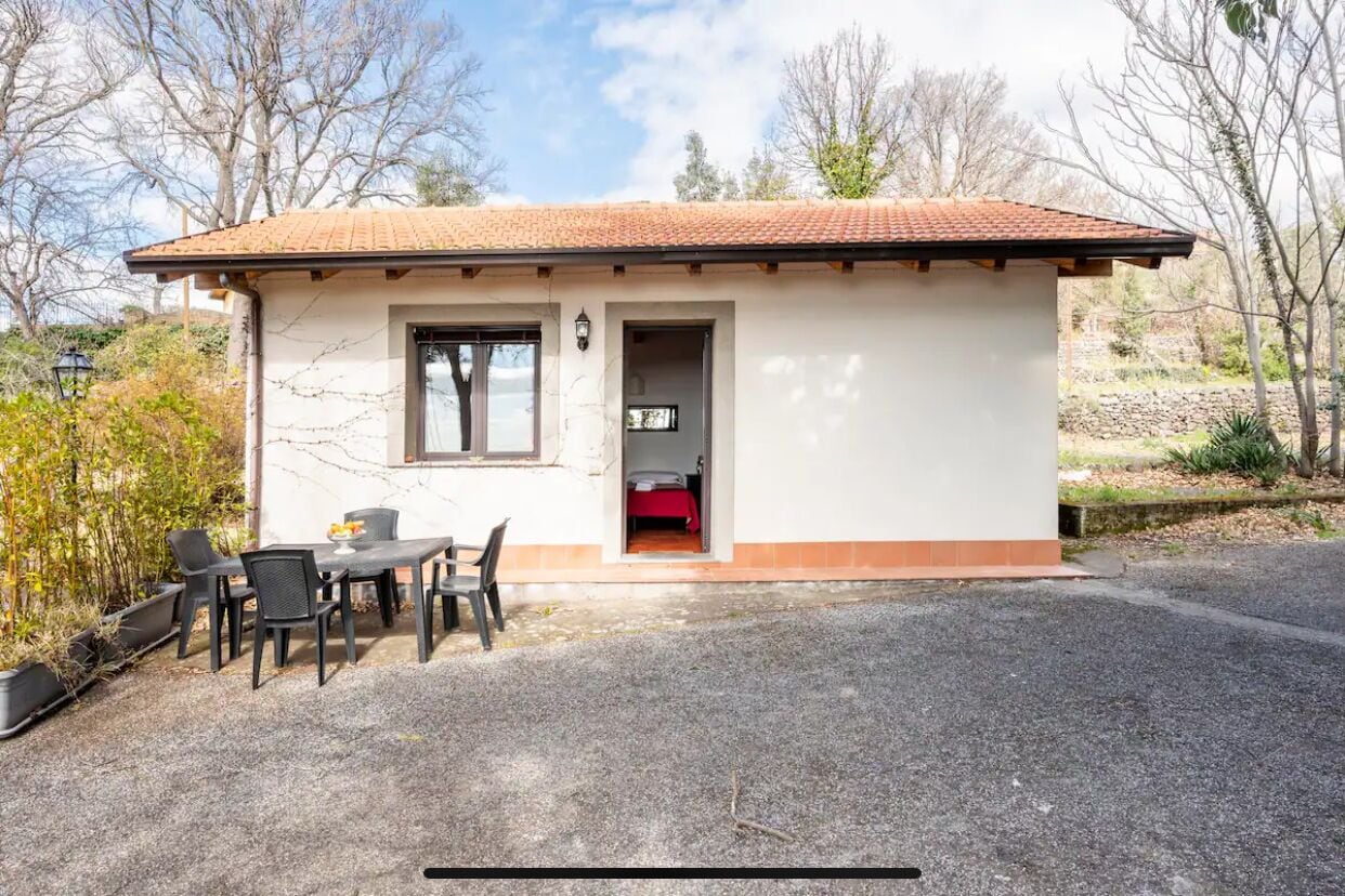 Een laag, eenlaags gebouw met een pannendak en een grindpad. Het heeft een eethoek met een tafel en stoelen, een klein raam en een deur die leidt naar een slaapkamer met een rode spreidek.'