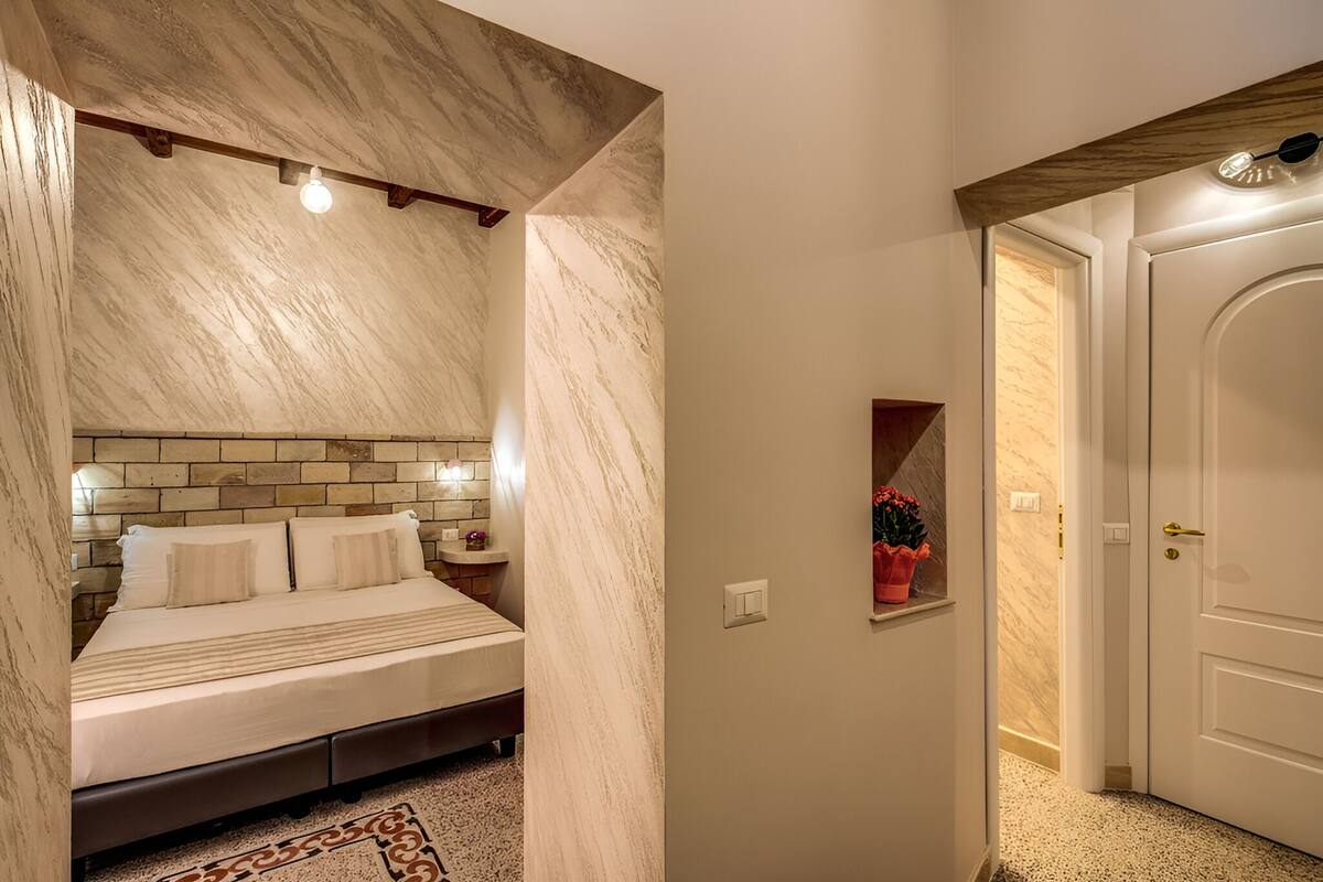 A bedroom with a marble headboard, a bed with white bedding, a nightstand, and a wall-mounted light fixture.