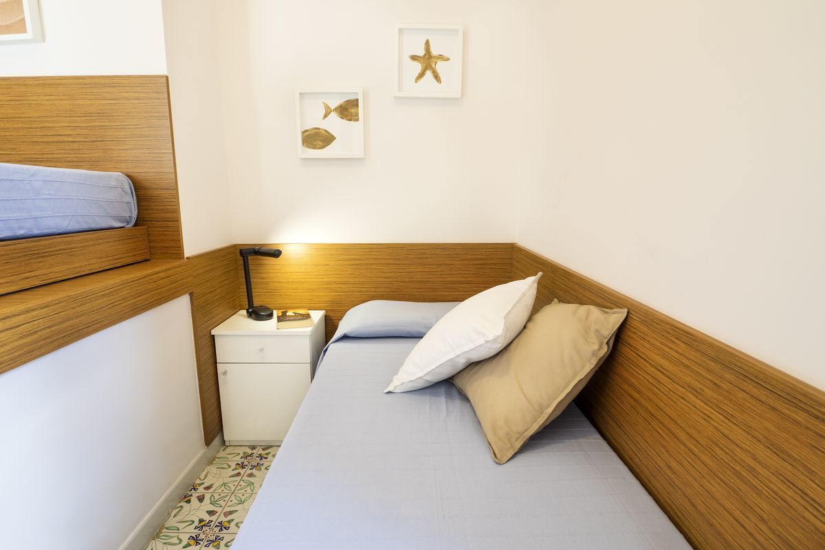 Una habitació petita i ben ordenada, amb un capçal de llit de fusta, un llit individual amb matalàs blau i almoades beix, una taula de nit amb una làmpada i una petita taula de nit blanca.