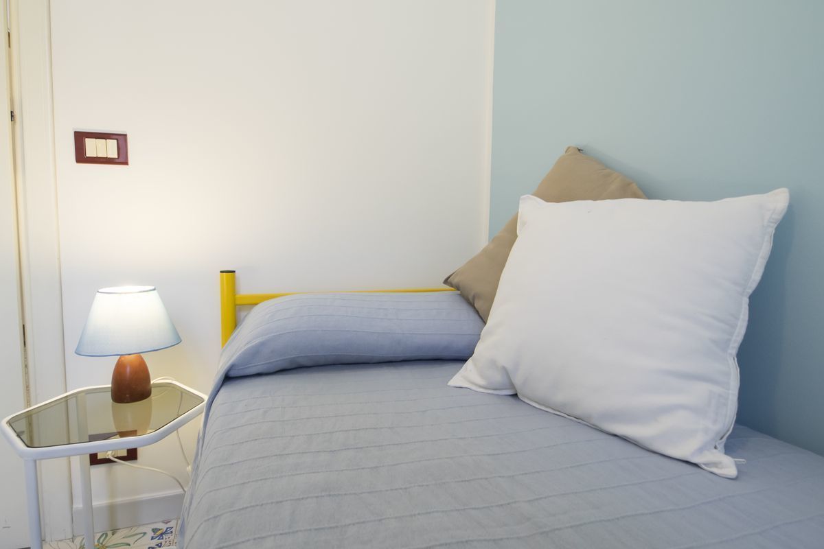 Un lit avec un oreiller blanc et un oreiller beige, une table de chevet avec une lampe et un interrupteur fixé au mur.