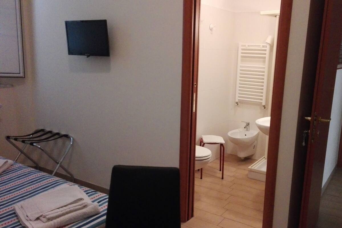 Une chambre d’hôtel équipée d’un lit, d’une chaise, d’un petit tabouret, d’une télévision, de toilettes et d’un lavabo.